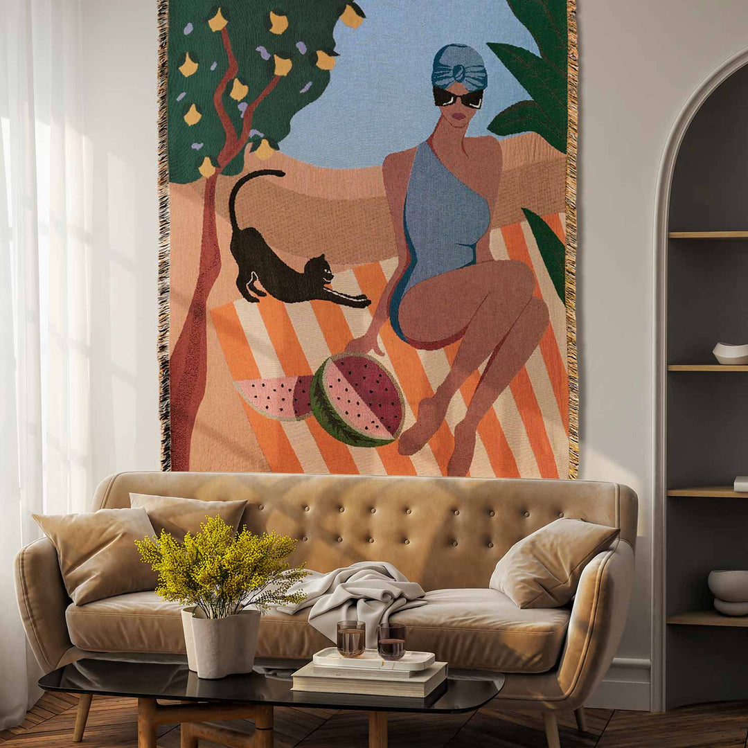 Atelier yuchi capri woven throw blanket with a scene featuring a woman in a blue bikini sitting by a pool with a black cat on her lap, surrounded by a watermelon, lemon tree, and striped background, draped over the back of a beige sofa.